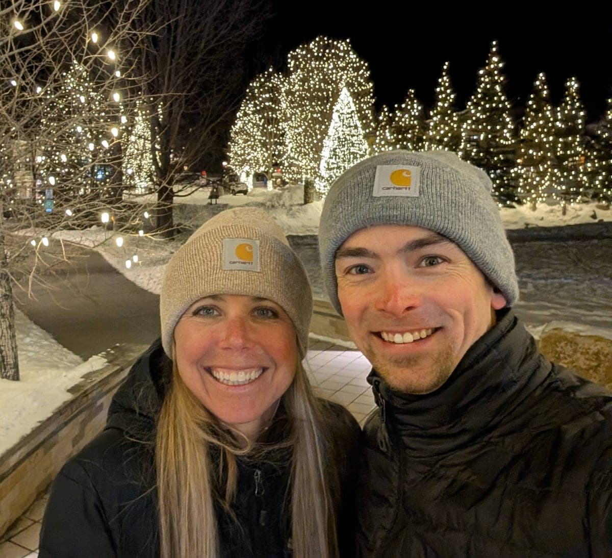 Andrew and Abby, the hosts of Lodestone Retreat, in winter hats in front of snowy trees strung with lights